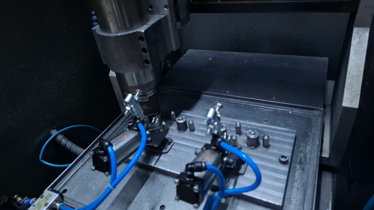 Detalhe interno de um centro de usinagem CNC mostrando o cabeçote e um dispositivo de fixação com grampos pneumáticos e mangueiras azuis